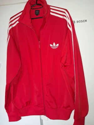Röd Adidas Originals track jacket storlek L - Röd track jacket från Adidas Originals med vita ränder längs ärmarna och klassisk trefoil-logga på bröstet. Dragkedja framtill, ribbade muddar och krage. Perfekt för en sportig och avslappnad stil. Storlek L