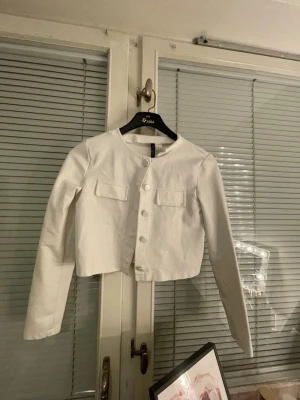 Vit cropped cardiganjacka H&M Elegant - Snygg vit cardiganjacka från H&M med rund halsringning och knäppning framtill. Jackan är cropped och har två dekorativa fickor på bröstet. Tillverkad i ett slätt tyg och har långa ärmar. Perfekt för en stilren look. Elegant
