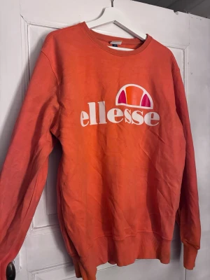 Orange sweatshirt från Ellesse - Säljer en orange sweatshirt från Ellesse med stort vitt och rosa logotryck på bröstet. Tröjan har rund halsringning, ribbade muddar vid ärmslut och nederkant samt en avslappnad passform. Perfekt för dig som gillar streetwear och sportig stil.