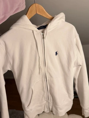 Vit zip hoodie från Polo Ralph Lauren - Klassisk vit hoodie med dragkedja från Polo Ralph Lauren. Har huva med dragsko, två fickor framtill och den ikoniska blå loggan broderad på bröstet. Tillverkad i mjukt bomullsmaterial för en skön och avslappnad look. Köpt från Zalando aldrig använt pågrund av fel storlek