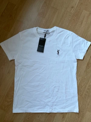 Vit t-shirt från Saint Laurent - Vit t-shirt från Saint Laurent med klassisk rund hals och svart YSL-logga broderad på bröstet. Tillverkad i mjuk bomull och har en stilren, enkel design som passar perfekt till jeans eller shorts. Helt oanvänd med lappen kvar, skriv vid frågor eller funderingar.