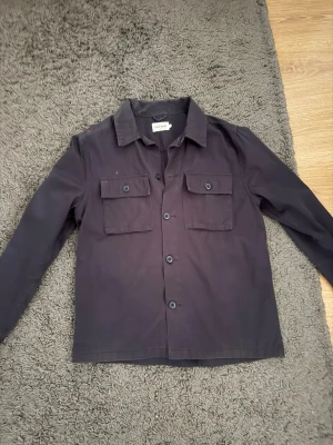 Mörkblå overshirt från Only & Sons - Stilren mörkblå overshirt från East & West med raka linjer, klassisk krage och två stora bröstfickor med lock och knapp. Tillverkad i ett mjukt bomullstyg och har knäppning framtill. Perfekt lager på lager-plagg för en avslappnad look.