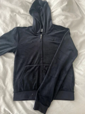 Mörkblå hoodie från Juicy Couture - Säljer en mörkblå hoodie i mjuk velour från Juicy Couture. Hoodien har dragkedja framtill, fickor och en broderad logga på bröstet. Perfekt för en chill och avslappnad stil. Klassisk huva och ribbade muddar vid ärmslut och nederkant.