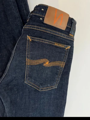 Mörkblå skinny jeans från Nudie - Säljer ett par mörkblå skinny jeans från Nudie Jeans Co med klassisk orange söm och vågdetalj på bakfickorna. Jeansen har läderpatch med logga i midjan och är tillverkade i denim av hög kvalitet. Perfekta för en stilren och trendig look.