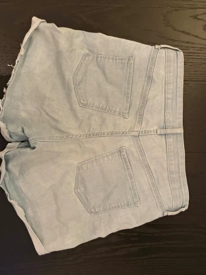 Ljusblå denimshorts med rå kant - Snygga ljusblå denimshorts med hög midja och rå, fransig kant nertill. Klassisk femficksmodell med bälteshällor och knappstängning framtill. Perfekta för varma dagar och enkel att matcha med allt.