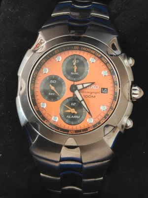 Seiko Chronograph  - Snygg Seiko kronografklocka i utmärkt skick med orange urtavla, silverfärgat armband i metall och flera subdials för minuter, sekunder och alarm. Nyligen fått ett färskt batteri. Klockan har tydliga indexmarkeringar, tachymeter och datumvisning. Kommer i originalask med extra länkar. Samt köp bevis sista foto visar tillverknings datum 