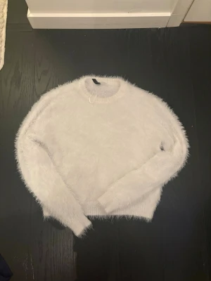 Vit fluffig stickad tröja - Supermjuk och fluffig vit stickad tröja med rund hals och långa ärmar. Tröjan har en mysig känsla och är perfekt för kalla dagar. Den har en lätt oversized passform och ribbade muddar vid ärmslut och nederkant.
