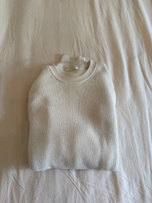 Zara Ribbstickad Tröja - Säljer en stilren vit ribbstickad tröja från Zara med rund hals och långa ärmar. Tröjan har breda muddar vid ärmslut och nederkant, vilket ger en snygg och modern look. Perfekt att matcha med jeans eller kjol för en clean outfit.
