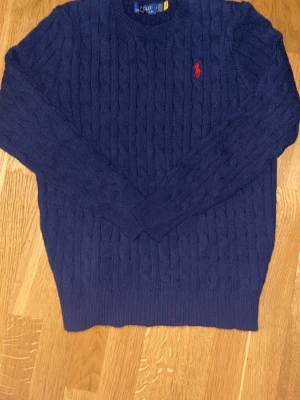 Mörkblå kabelstickad tröja Polo Ralph Lauren - Säljer en mörkblå kabelstickad tröja från Polo Ralph Lauren med rund hals och lång ärm. Tröjan har ett klassiskt rött broderat logotyp på bröstet och ribbade muddar vid ärmslut och nederkant. Perfekt för dig som gillar stilrena och tidlösa plagg. Den är lite tightare på ärmanarna och mindre än M. Den passar ca 12-14 åring.