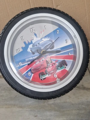 Väggklocka - Säljer en rund väggklocka med motiv från Disney Cars och ram som ser ut som ett bildäck. Har legat i förrådet och kan därför behövas en puttsning. Perfekt för dig som gillar bilar och cool inredning.