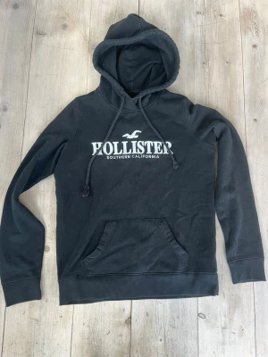 Svart hoodie från Hollister - Svart hoodie från Hollister med vit logga och texten 'Southern California' på bröstet. Klassisk känguruficka framtill, ribbade muddar och dragsko i huvan. Perfekt för en chill och avslappnad stil. Passar dig som är ca 165-170 cm lång.