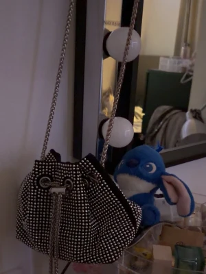 Svart bucket bag med silverdetaljer - Cool svart bucket bag med massor av små silvriga nitar och kedjerem. Väskan har dragskostängning och en trendig form som sticker ut. Perfekt för dig som vill ha en unik accessoar med lite edge. Materialet känns som textil med metalliska detaljer.Denna väskan köpte jag för ca 2 år sedan på västkusten.