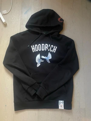 Svart HOODRICH hoodie med fågeltryck - Svart hoodie från HOODRICH med vit logga och två vita fåglar tryckta på bröstet. Klassisk huva med dragsko och känguruficka framtill. Liten etikett nertill med HOODRICH-logga och fågelmotiv. Perfekt för en streetwear-look.