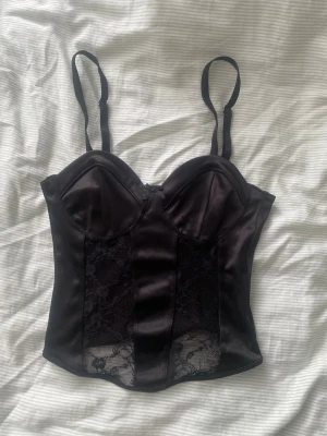 Svart satin korsett med spetsdetaljer - Snygg svart korsett i satin med genomskinliga spetspaneler framtill och justerbara axelband. Kuporna är formade för extra stöd och en feminin siluett. Perfekt för att lyfta din outfit med en elegant touch.