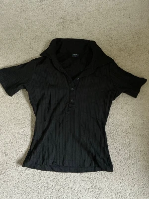 Svart ribbad polotopp från Filippa k  - Svart ribbad polotopp från Filippa K med korta ärmar och knappar framtill. Toppen har en hög krage som kan vikas ner och en figurnära passform. Perfekt för dig som gillar en stilren och enkel look.