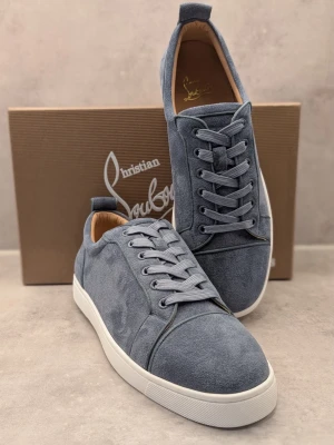 Blå mockasneakers från Louboutin - Snygga blå sneakers i mocka från Christian Louboutin med klassisk låg profil och vit sula. Skorna har den ikoniska röda undersulan, ton-i-ton snörning och diskreta sömdetaljer. Perfekta för dig som vill ha en stilren och exklusiv look.