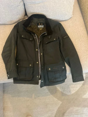 Grön Barbour jacka med fickor - Klassisk mörkgrön jacka från Barbour med flera stora fickor framtill, krage i kontrastmaterial och dragkedja samt knappar. Perfekt för höst och vår, med tidlös design och praktiska detaljer. Storlek S