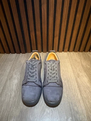 Gråa Louboutins med röd sula - Snygga grå Louboutins i mockaliknande material med klassisk låg profil och matchande grå skosnören. Insidan är beige och yttersulan är riktigt cool med röd detalj undertill. Perfekta för dig som vill ha en clean look med en twist.