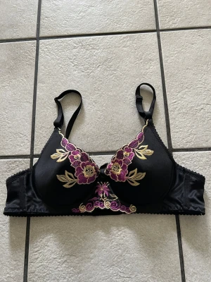 Svart bh med broderade blommor storlek 70 A/B - Snygg svart bh med vackra broderade blommor i lila och guld på kuporna. Justerbara axelband och hakknäppning bak. Bh:n har en feminin design med fina detaljer och mjukt material som känns bekvämt mot huden.