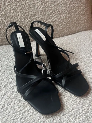 Svarta klackar från H&M - Stilrena svarta sandaler från H&M med tunna remmar och fyrkantig tå. Skorna har en smal klack och justerbar rem runt hälen. Tillverkade i läder och har en minimalistisk och trendig design som lyfter varje outfit. Klackarna är helt oanvända och jag säljer dem då dem är för små för mig! 