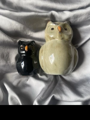 Keramiska ugglor prydnadsfigurer - Två keramiska ugglor i olika storlekar, en i beige och en i svart. Båda har orange näbb och detaljerade ögon. Perfekta som dekoration i hemmet eller som samlarobjekt. Säljes tillsammans.