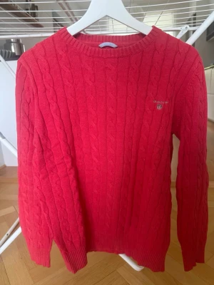 Röd kabelstickad tröja från Gant - Snygg röd kabelstickad tröja från Gant med rund halsringning och diskret logga broderad på bröstet. Tröjan har långa ärmar och ribbade muddar vid ärmslut och nederkant. Perfekt för dig som gillar klassisk och stilren design.