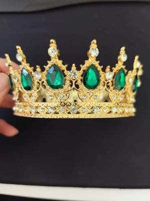 Guldig krona med gröna stenar - Snygg och detaljerad krona i guldfärgad metall med stora gröna droppformade stenar och klara kristaller. Perfekt för maskerad, cosplay eller temafest där du vill sticka ut som kunglighet. Kronan har en klassisk form och gnistrar rejält i ljuset.