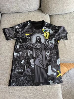 Nike, Brasilien fotbollströja - Snygg svart t-shirt från Nike med Brasilien-tryck och unikt grafiskt mönster framtill. T-shirten har korta ärmar, rund hals och detaljer i gult. Perfekt för en sportig look. Märket på baksidan vid nacken har lossnat med användning.
