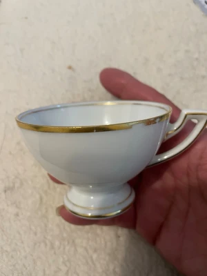 Vit kopp med guldkant i porslin - Elegant vit kopp i porslin med guldfärgad kant och dekorativt handtag. Koppens form är klassisk med rundad botten och fot. Perfekt för te eller kaffe och ger en lyxig känsla till fikastunden.