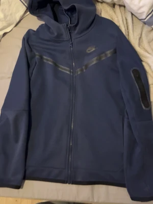 Mörkblå Nike Tech Fleece hoodie - Nike Tech Fleece hoodie i mörkblått med hel dragkedja och svarta detaljer över bröstet. Inga hål eller något liknande Har huva och en ficka med dragkedja på ärmen. Perfekt för en sportig och stilren look. 
