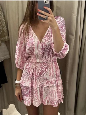 Rosa och vit kortklänning med volang - Söt kortklänning i rosa och vitt med blommigt mönster, v-ringning och volangdetaljer. Klänningen har trekvartsärm och resår i midjan för en bekväm passform. Perfekt för varma dagar.