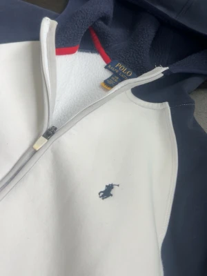 Vit och marinblå hoodie från Polo Ralph Lauren - Snygg hoodie från Polo Ralph Lauren i vitt med marinblå ärmar och huva. Dragkedja framtill, två fickor med dragkedja och klassisk Polo-logga broderad på bröstet. Mjuk fleece på insidan och röd detalj vid halsen. Perfekt för en avslappnad stil. 
