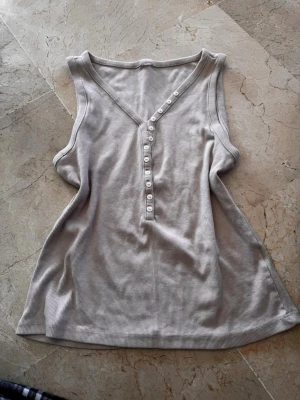 Ribbad beige linnetopp med knappar - Snygg ribbad linnetopp i beige med v-ringning och knappar framtill. Ärmlös modell som passar perfekt till varma dagar. Enkel och stilren design som är lätt att matcha med olika outfits.