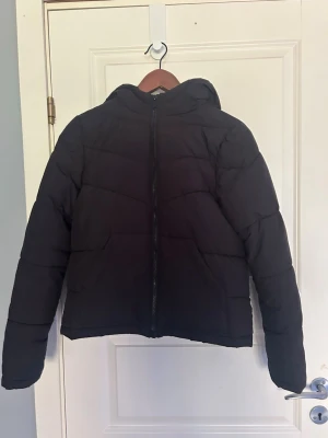 Svart pufferjacka - Säljer en svart pufferjacka med huva och dragkedja framtill. Jackan har en klassisk quiltad design och är perfekt för kalla dagar. Den är vadderad och har en rak passform som ger en stilren look. Materialet är skönt och jackan är lätt att matcha med olika outfits. Perfekt för en kallare höst/vår dag!
