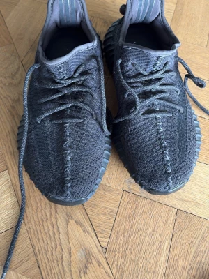 Svarta Yeezy Boost 350 V2 sneakers - Säljer ett par svarta Yeezy Boost 350 V2 sneakers med stickad ovandel, ribbad sula och röda detaljer på hälen. Skorna har en modern siluett, snörning och är tillverkade i textil och syntetmaterial. 