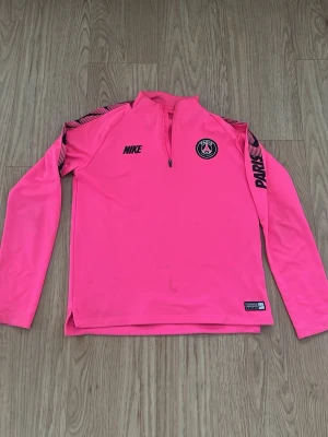 Rosa Nike PSG träningströja - Säljer en snygg rosa långärmad träningströja från Nike och Paris Saint-Germain. Tröjan har halv dragkedja, svarta detaljer på axlarna och PSG-logga på bröstet. Perfekt för fotbollsträning eller sportiga dagar. Den är ganska använd fast om man tvättar rätt så går allt bort