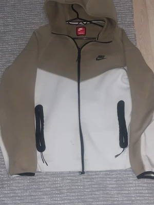 Nike beige och vit hoodie med dragkedja - Nike hoodie i beige och vitt med svart dragkedja och logga på bröstet. Utrustad med huva och två svarta fickor framtill. Tillverkad i mjukt material, perfekt för en avslappnad streetwear-look. 1400 kr ny pris 