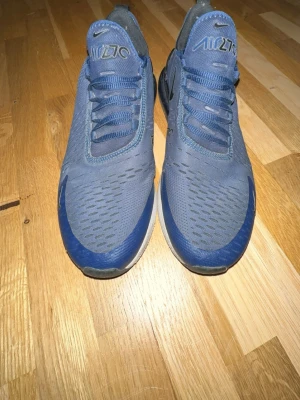 Nike Air Max 270 blå sneakers - Snygga Nike Air Max 270 sneakers i blått med svarta detaljer och vit sula. Ovandelen är i mesh för bra ventilation och skon har en tydlig Air Max-dämpning i hälen. Klassisk Nike-logga på sidan och tungan. Perfekta för dig som gillar sportig stil.