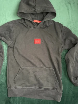 Svart hoodie från HUGO med röd patch - Snygg svart hoodie från HUGO med en tydlig röd patch med logga på bröstet. Hoodien har huva, känguruficka och ribbade muddar vid ärmslut och nederkant. Perfekt för en avslappnad och trendig look. Tillverkad i mjuk bomullsblandning. Pris kan diskuteras. MVH stentorparchive⚓️