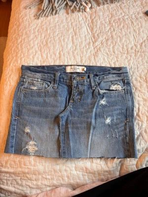 Blå jeanskjol med slitningar - Snygg blå jeanskjol från Abercrombie & fitch med slitna detaljer och rå kant nertill. Klassisk femficksmodell med bälteshällor och knappstängning. Perfekt för dig som gillar en avslappnad och trendig stil.