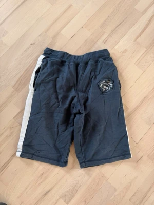Mörkblå sportiga shorts med vit rand - Mörkblå shorts med vit rand längs sidorna och ett tryckt emblem på vänster ben. Resår i midjan och två sidofickor. Perfekta för en avslappnad och sportig stil.