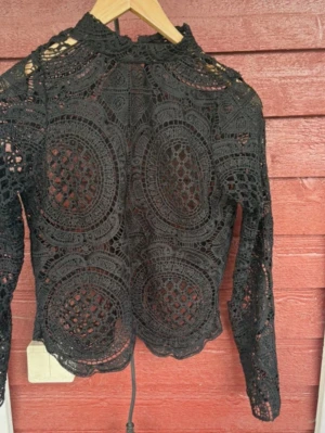 Svart spetsblus från BikBok - Snygg svart spetsblus från BikBok med långärmad design och hög hals. Blusen har ett detaljerat virkat mönster och är genombruten vilket ger en cool och trendig look. Perfekt att styla med jeans eller kjol för en extra touch.