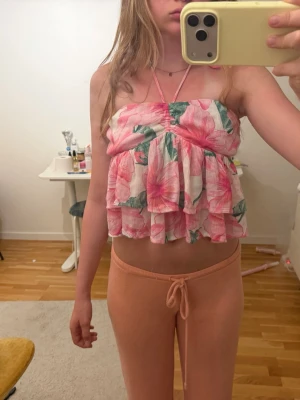 Blommig volang bandeau topp - Söt bandeau topp med volanger och blommigt mönster i rosa och grönt. Toppen är axelbandslös med en halterneck-detalj och har en luftig, somrig känsla. Perfekt för dig som gillar färgglada och lekfulla plagg.