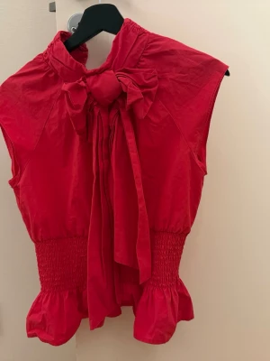 Röd ärmlös topp med rosett - Snygg röd ärmlös topp med stor rosett vid halsen och smockad midja. Toppen har volangdetaljer nertill och är gjord i ett lätt tyg som känns bekvämt. Perfekt för att lyfta din outfit med en färgklick och cool design. Den är lite rosa röd!