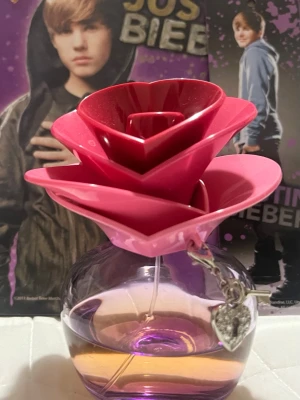 Someday parfym av Justin Bieber - En söt parfymflaska från Justin Bieber med en unik blomformad kork i olika rosa nyanser. Flaskan är rund och genomskinlig med lila toner och har ett hjärtformat silverhänge. Perfekt för dig som gillar snygg design och popkultur.