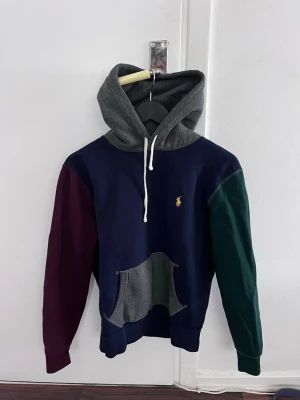 Flerfärgad hoodie från Polo Ralph Lauren - Snygg hoodie från Polo Ralph Lauren i flera färger: marinblå kropp, vinröd ärm, grön ärm och grå huva. Klassisk känguruficka framtill och vit dragsko. Liten gul broderad logga på bröstet. Perfekt för dig som gillar streetwear och färgkombos.