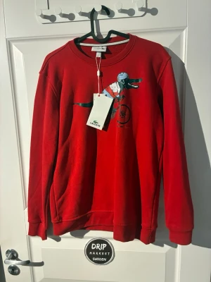 Röd sweatshirt Lacoste krokodiltryck - Röd sweatshirt från Lacoste med ett coolt tryck av en krokodil på cykel med hjälm. Tröjan har rund halsringning, ribbade muddar och är tillverkad i mjukt material. Perfekt för dig som vill sticka ut med en unik och lekfull design.