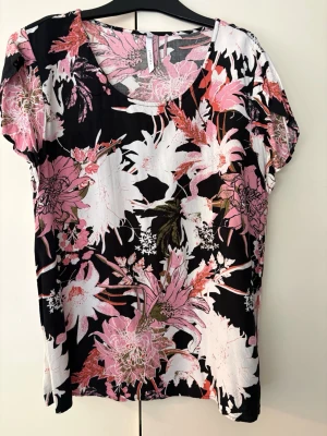 Blommig topp från Jyothi strl.XL - Snygg topp från Jyothi med stort blommigt mönster i rosa, vitt och grönt på svart botten. Toppen har korta ärmar och rund halsringning. Perfekt för dig som gillar färg och mönster!