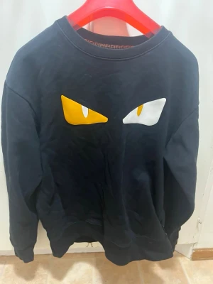 Fendi sweatshirt - Cool svart sweatshirt från Fendi med ikoniskt ögonmotiv i gult och vitt på bröstet. Rund halsringning med Fendi-logga på insidan av kragen. Perfekt statement-plagg för dig som gillar streetwear och designer.