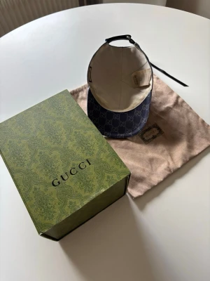 Blå keps från Gucci med monogram - Säljer en snygg blå keps från Gucci med det klassiska GG-monogrammet över hela kepsen. Kepsen har böjd skärm, justerbart band bak och kommer med originalförpackning och dustbag. Materialet är canvas med insida i ljust tyg. Perfekt för dig som gillar exklusiva accessoarer.